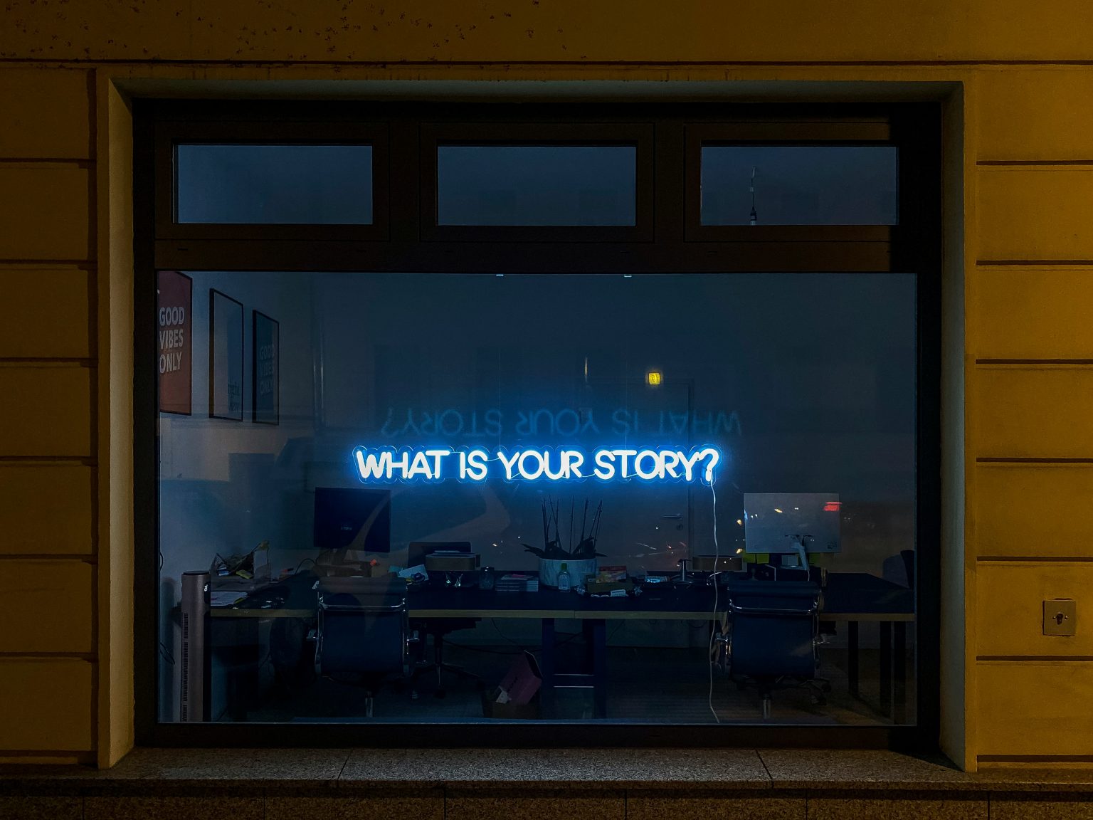Enseigne néon bleue dans une vitrine de bureau sombre affichant la question : "WHAT IS YOUR STORY?" (Quelle est votre histoire ?).